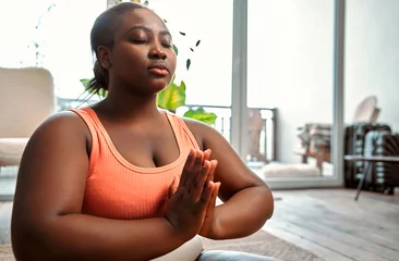 Tuinposter Relaxing yoga. Serene curvy woman in sport attire clasping hands in namaste and keeping eyes closed during meditation. African female sitting in lotus pose at living room and feeling peace of mind. © HBS
