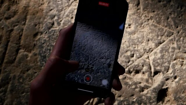 A man's hand holding a smartphone photographs an ancient image of the star of Israel on a stone. A man uses a mobile phone camera to film the Star of David carved on an ancient stone slab.