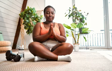  Yoga for health. Overweight afro woman meditating in lotus pose with closed eyes and namaste gesture. Relaxed young female in sport clothes sitting on floor near dumbbells and water bottle. © HBS