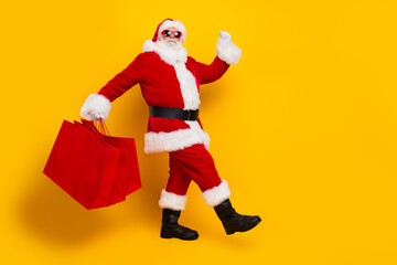 Fototapeta na wymiar Side shot of walk shopaholic santa claus celebrate holly jolly happy new year bring red bargains bags isolated on yellow color background