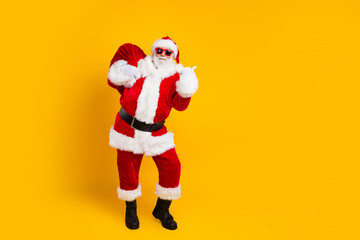 Fototapeta na wymiar Full size portrait of eccentric santa carry christmas presents sack indicate finger empty space isolated on yellow color background