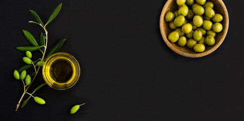 Olive oil in a glass bowl, branch with green olive and green olive in the bowl on the black background. Top view. Copy space.