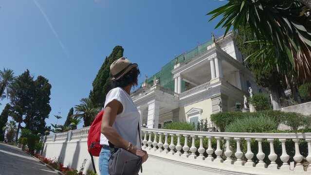 Tourist woman with hat and backpack sightseeing in the garden in Achilleion Palace, in Corfu island, Greece, slow motion