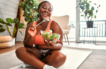 Healthy diet and sport. Beautiful plump woman in sport clothes eating vegetable salad from glass bowl while sitting on floor. Black young female following slimming and exercising program at home. - obrazy, fototapety, plakaty