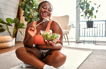 Poster Healthy diet and sport. Beautiful plump woman in sport clothes eating vegetable salad from glass bowl while sitting on floor. Black young female following slimming and exercising program at home. © HBS