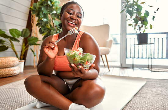 Healthy diet and sport. Beautiful plump woman in sport clothes eating vegetable salad from glass bowl while sitting on floor. Black young female following slimming and exercising program at home.