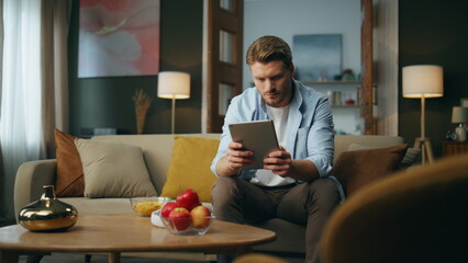 Calm man browsing tab device at home. Serious guy sitting sofa scrolling tablet