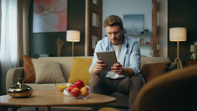 Calm man browsing tab device at home. Serious guy sitting sofa scrolling tablet