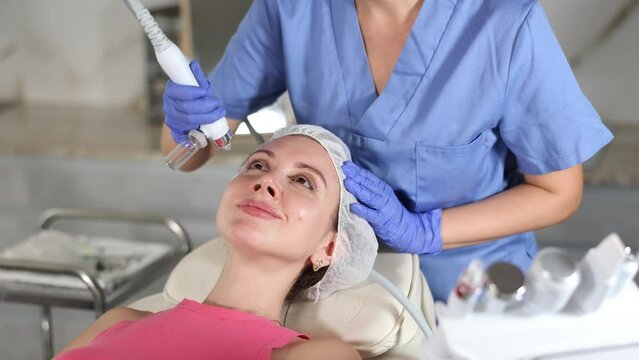 Young female cosmetologist performs hardware facial moisturizing procedure to adult female patient