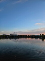 a river with trees, water, sky and ferris wheel in the background