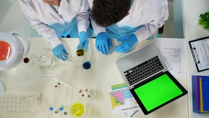 There is a close up of a man and a woman in the frame above. They are scientists, the woman holds a flask with a liquid inside. The man is typing on a laptop with a green screen. A place to advertise
