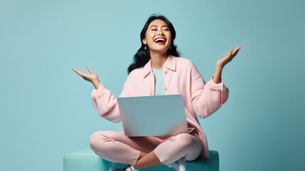 Excited young woman raising her fists in the air with a joyful expression, likely celebrating success while working on her laptop against a blue background.