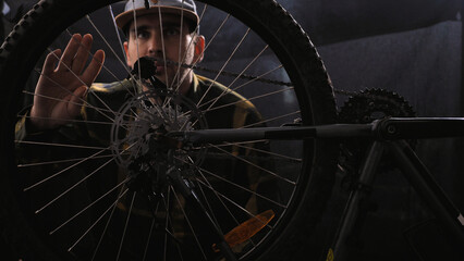 A bike mechanic wearing a baseball cap and shirt repairs a bicycle in a workshop. A man spins a...