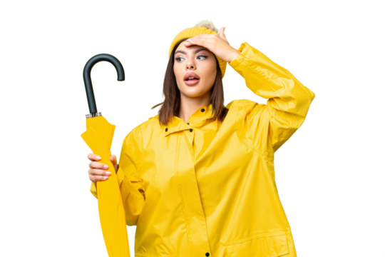 Young Russian woman with rainproof coat and umbrella over isolated chroma key background doing surprise gesture while looking to the side