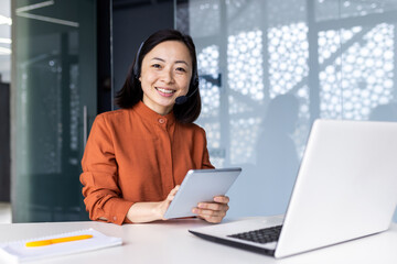 Portrait of successful satisfied female online customer service support worker, Asian business woman looking at camera, holding tablet computer, using headset and laptop for video call.