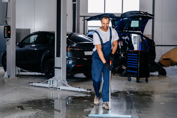 Arabian service staff man using a mop to remove water in the uniform cleaning the protective clothing of the new epoxy floor in an empty warehouse or car service center. - obrazy, fototapety, plakaty