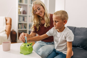 Happy mother is showing her son how to put money in a piggy bank and save it.
