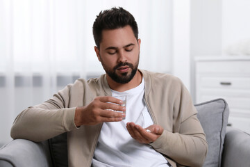 Man with glass of water taking antidepressant pill in armchair at home