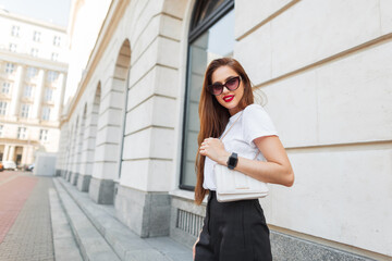 Successful happy business lady in fashion outfit with sunglasses with a white stylish bag walking in the city near a vintage building