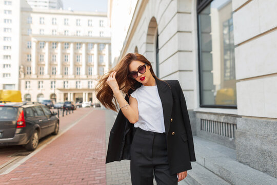 Chic young urban fashionable business beauty woman with sunglasses in trendy fashion black casual clothes with blazer walks in the city