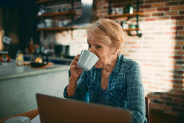 Senior woman drinking coffee looking at laptop in kitchen