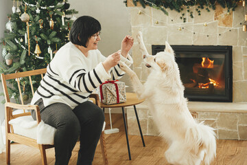 Happy senior woman playing with adorable white dog in stylish festive christmas living room. Beautiful woman enjoying cozy fireplace with cute pet on background of decorated christmas tree