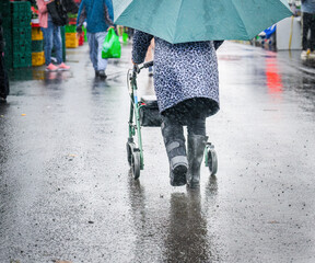 Woman wearing moon boot, pushing a mobility walker in the rain. Unrecognizable people shopping in...