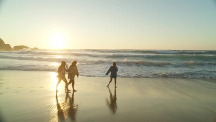 Cinematic sunset vibes in this moody warm summer shot of three happy women run away from ocean waves on sandy beach in California. 