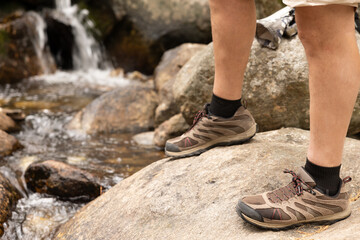 legs of man in hiking boots trekking over rocks with river and waterfall in background