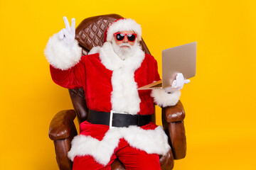 Photo of old santa claus xmas party holding laptop order gifts in internet for 1 second showing v sign isolated on yellow color background