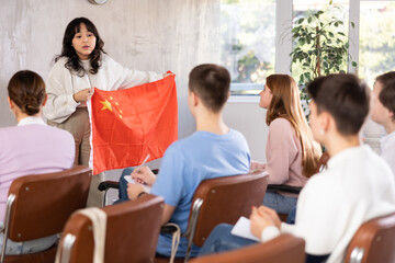 Focused young Asian girl holding national flag of China and talking about history of country in front of group of coursemates during college class