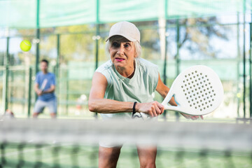 Concentrated elderly woman padel player hitting ball with a racket on a hard court
