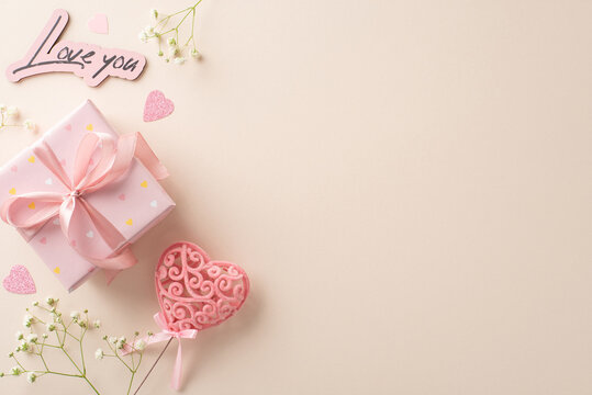 Love Celebration: Top view image featuring wrapped gift, gypsophila, heart confetti, love declaration, and heart decor on stick. Pastel beige background with text or advertising space