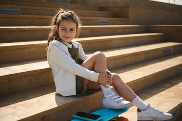 Caucasian little girl with skateboard and smart mobile phone, sitting on steps outdoors and smiling looking at camera