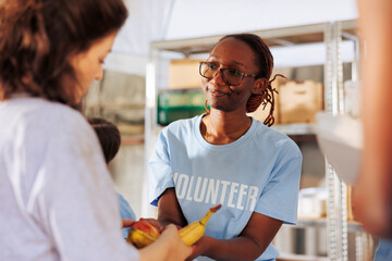 Detailed image showing african american female volunteer offering free meals to the poor and needy. Nice black woman at an outdoor food bank sharing fresh produce and nourishments to underprivileged. - obrazy, fototapety, plakaty