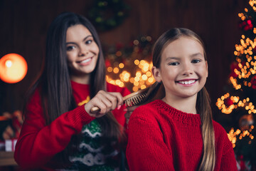 Photo of pretty charming little siblings dressed red sweaters combing hair doing x-mas hairdo indoors christmas apartment home