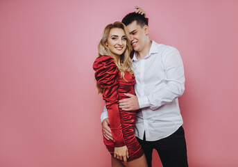 A stylish young man and a beautiful smiling woman in a red dress are hugging on a pink background...