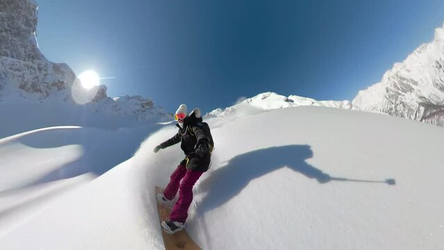 SELFIE, LENS FLARE: Happy woman enjoys snowboarding untouched snowy terrain on an adventure heliboarding trip in beautiful Albanian Alps. She has fun riding and spraying clouds of fresh powder snow.