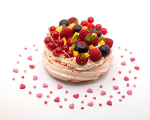 Meringue pavlova dessert, decorated with whipped cream,  berries and mango. Surrounded with heart shaped sprinkles. Photographed against a white background.