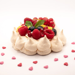Meringue pavlova dessert, decorated with whipped cream,  berries and mango. Surrounded with heart shaped sprinkles. Photographed against a white background.