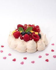 Meringue pavlova dessert, decorated with whipped cream,  berries and mango. Surrounded with heart shaped sprinkles. Photographed against a white background.