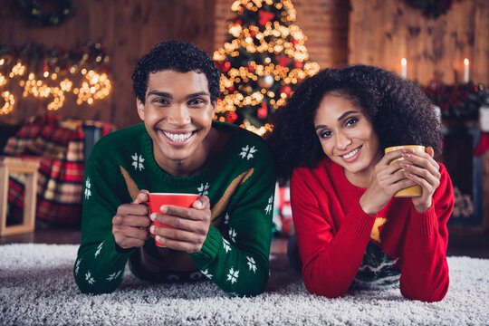 Portrait of two funny cheerful people lying carpet floor hold hot chocolate mug new year tree lights apartment indoors