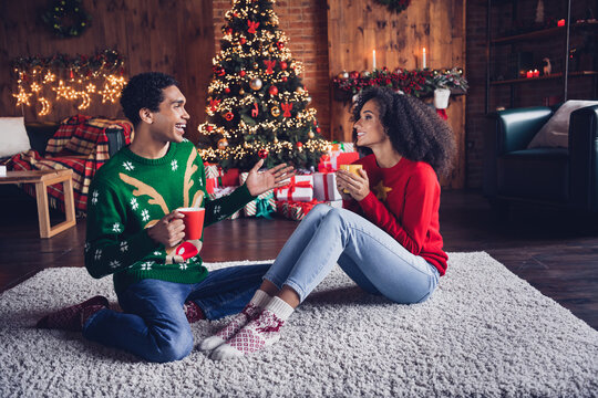 Full size portrait of two funny positive people sit carpet floor drink hot chocolate speak new year tree lights indoors