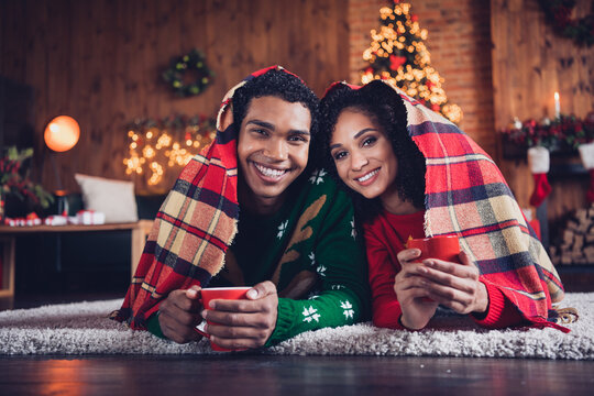 Portrait of two cheerful people lying carpet floor covered blanket hold tea mug new year tree lights apartment indoors