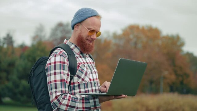 Stylish red-bearded man with backpack, in yellow sunglasses touches beard thoughtfully, points finger up, exclaiming. Blurred background of autumn trees. High quality 4k footage