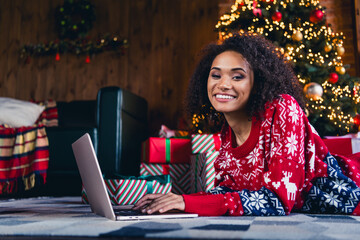 Photo of charming black skin girl lying on fluffy carpet chatting with friends on macbook xmas day...