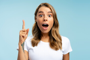 Amazed student holding finger up having creative idea isolated on blue background, education concept.  Beautiful emotional blonde woman with open mouth pointing hand looking at camera in studio