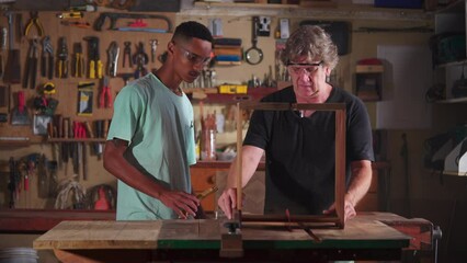 Carpenter working at workshop with the help of apprentice, small business worker doing woodwork in carpentry workplace