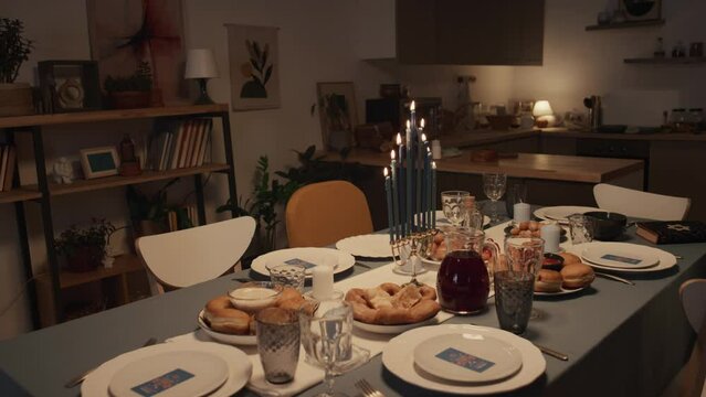 Medium full shot of festive table laid out for Hanukkah dinner at family home - menorah with burning candles, holy Torah book, plates with latkes and sufganiyot, waiting for arrival of guests