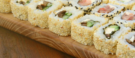 Close-up of a lot of sushi rolls with different fillings lie on a wooden surface. Macro shot of cooked classic Japanese food with a copy space.